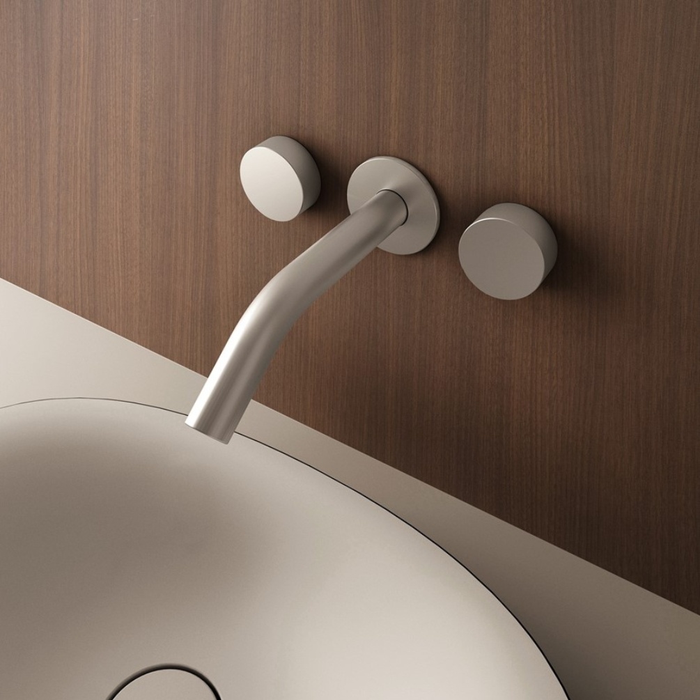 Lifestyle image of the Crosswater Ixora Stainless Steel 3 Tap Hole Wall Mounted Basin Mixer attached to a wood-effect bathroom wall, above a countertop basin